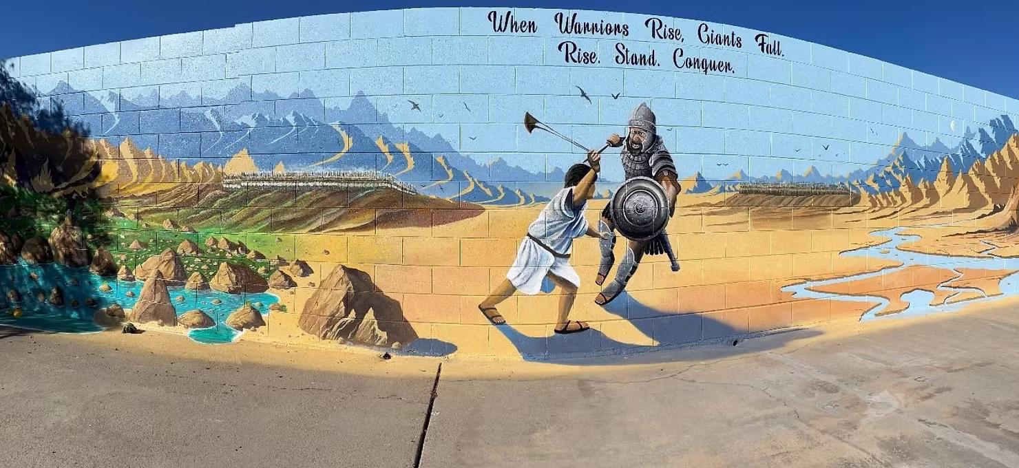 a wall with a mural of david fighting goliath. With text above it that says "when warriors rise, giants fall. rise. stand. conquer."