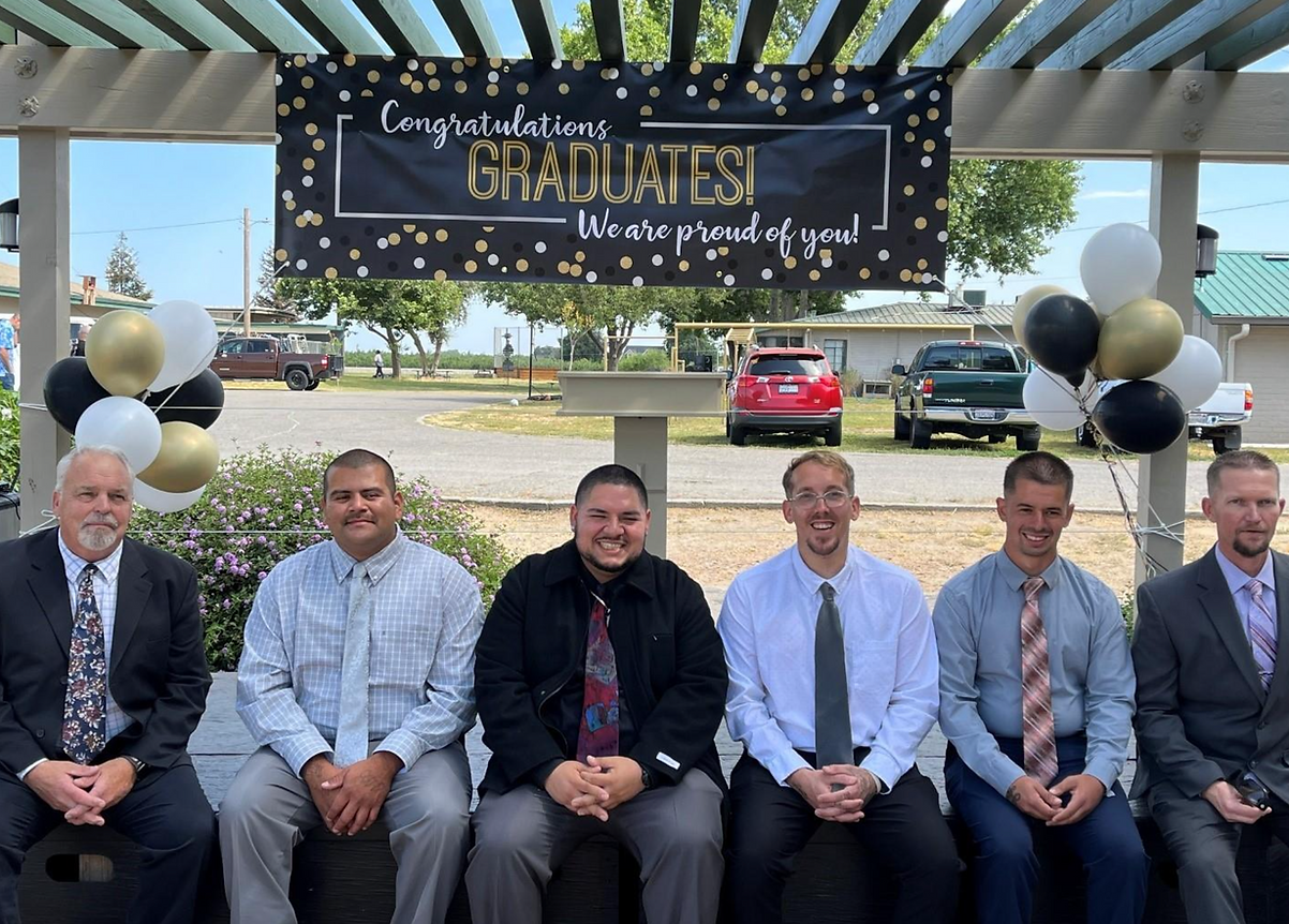 six men sitting on a bench smiling below a "congratulations Graduates! we are proud of you!" sign