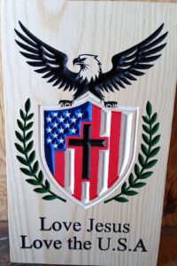 wood sign with a bald eagle holding a shield with an american flag and cross inside it. Vines are on either side of the shield. There is text under the shield that says "Love Jesus, Love the USA"