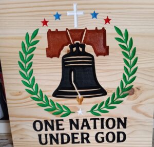 wood sign with the liberty bell in the middle with vines, stars, and a cross surrounding it in a circle. Also has text that says "One nation under God" at the bottom