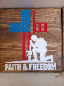 wood sign with an american flag shaped like a cross and a soldier kneeling down next to it. Below that it says, "Faith & Freedom"