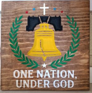 wood sign with the liberty bell in the middle with vines, stars, and a cross surrounding it in a circle. Also has text that says "One nation under God" at the bottom