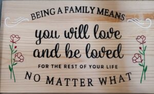 wood sign with flowers on either side of text. Test says, "Being a family means you will love and be loved for the rest of your life no matter what"