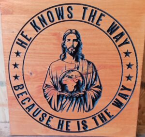 Wood sign with Jesus in the middle holding the world in his hands. A circle surrounding him with the text "He knows the way because he is the way" in it