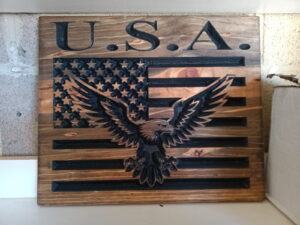 wood sign with american flag in the background and a bald eagle with its wings spread in the foreground. Also has the text "USA" above the flag/eagle
