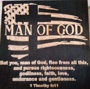 wood sign with "man of God" written in the middle of a waving american flag and cross. Below that is text saying "But you, man of God, flee from all this, and pursue righteousness, godliness, faith, love, endurance and gentleness. 1 Timothy 6:11"