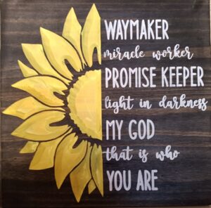 wood sign with half a yellow sunflower on the left side and white text on the right side that says "Way maker, miracle worker promise keeper light in the darkness my God that is who you are"