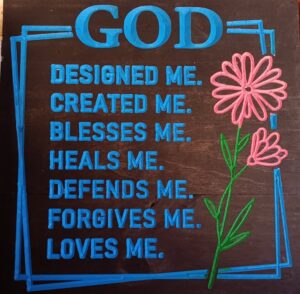 wood sign with blue text that says "God designed me. Created me. Blesses me. Heals me. Defends me. Forgives me. Loves me." Text is surrounded by blue box. Also have green and pink flowers on the right side of the text