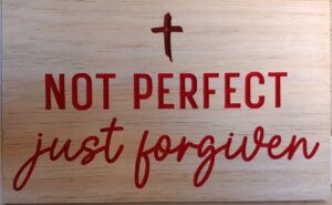 wood sign with a red cross at the top and red text below it that says "Not perfect just forgiven"