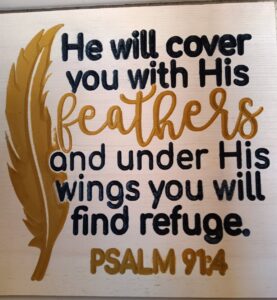 wood sign with gold feather on the left and black and gold text that says "He will cover you with His feathers and under his wings you will find refuge. Psalm 91:4"