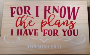 wood sign with red and pink text that says "For I know the plans I have for you" with a white swirly line below and "Jeremiah 29:11" written in white at the bottom.