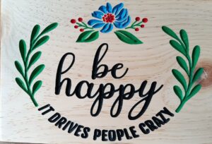 wood sign with vines and a flower and text that says: by happy, it drives people crazy"