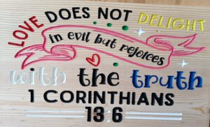 wood sign with colorful text that says "Love does not delight in evil but rejoices with the truth. 1 Corinthians 13:6"